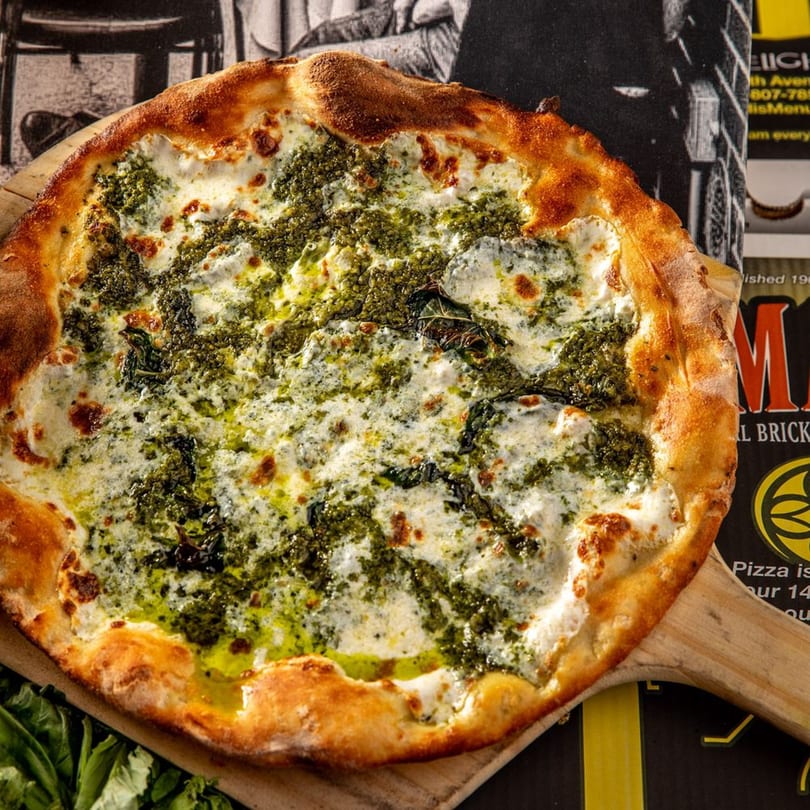 A Brooklyn Born pesto pizza with white cheese and a golden crust on a wooden peel.