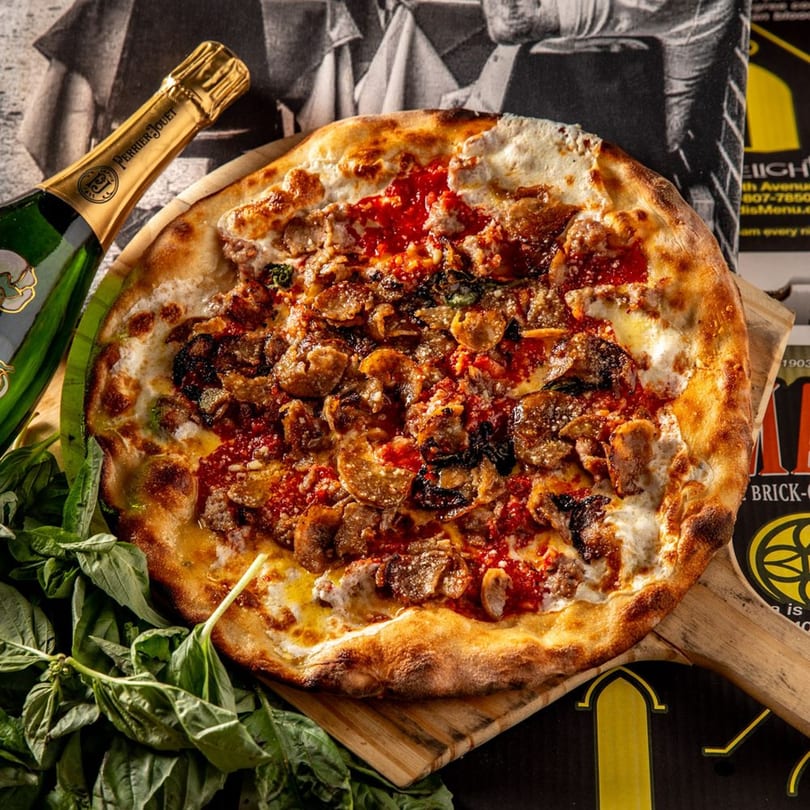Brooklyn Born pizza with sausage, mushrooms, and red sauce, next to a bottle of Perrier-Jouët champagne.