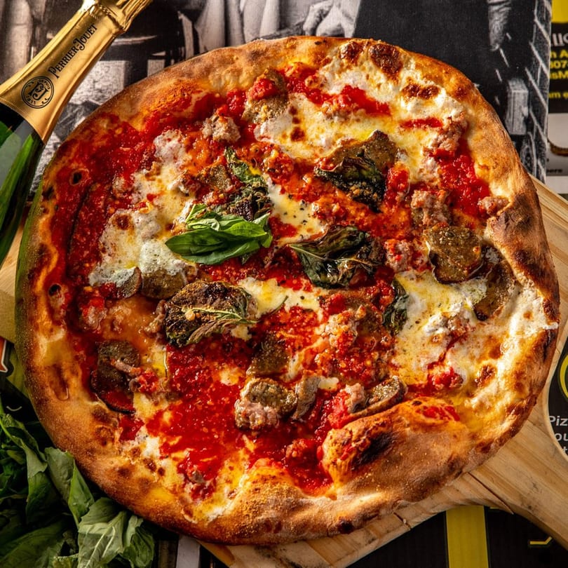 A Brooklyn Born pizza with sausage, mushrooms, basil, and tomato sauce, next to a champagne bottle.