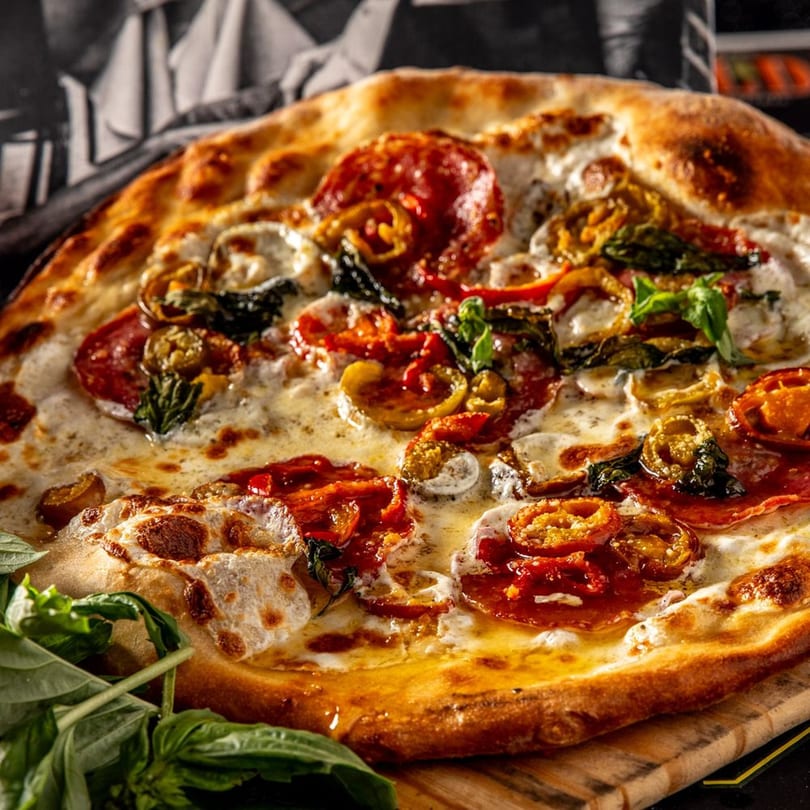 A Brooklyn Born pizza with pepperoni, melted cheese, basil, and chili peppers on a wooden board.